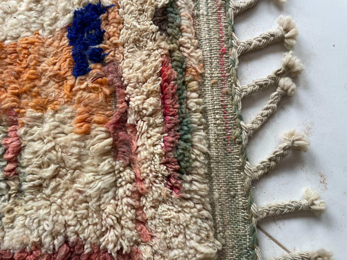 Detail view of a vintage Moroccan rug featuring white, orange, blue, and red colors with fringe edges.