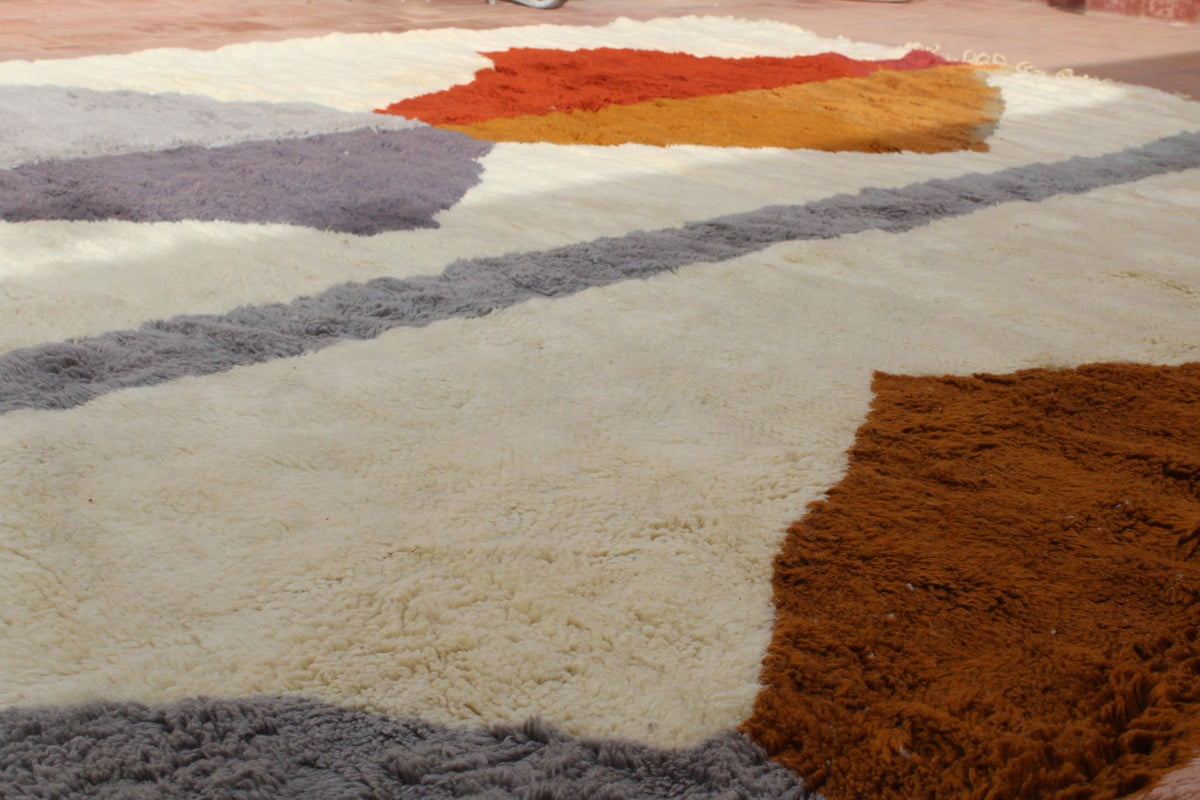 Lifestyle shot of a Moroccan rug with geometric patterns in beige, orange, and purple tones on a textured surface.