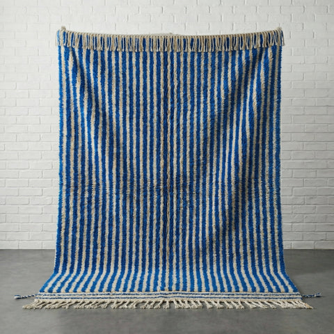 Front view of a blue striped Moroccan rug with fringe edges in a studio setting.