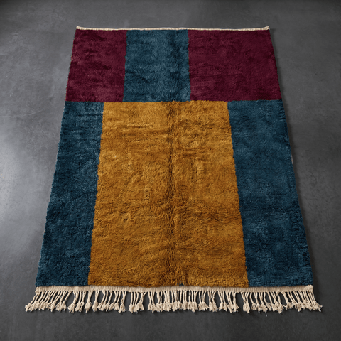 Top view of a handmade Moroccan rug showcasing a geometric pattern in burgundy, teal, and gold with fringe edges.