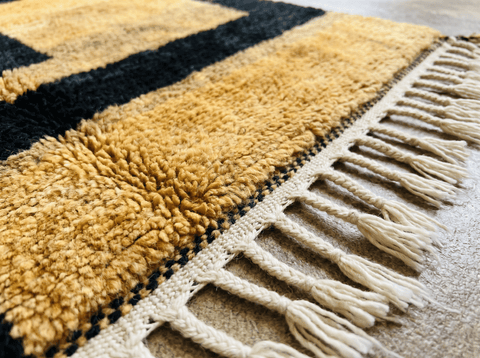 Detail view of a golden yellow rug with fringe edges and a textured pattern in a natural setting.