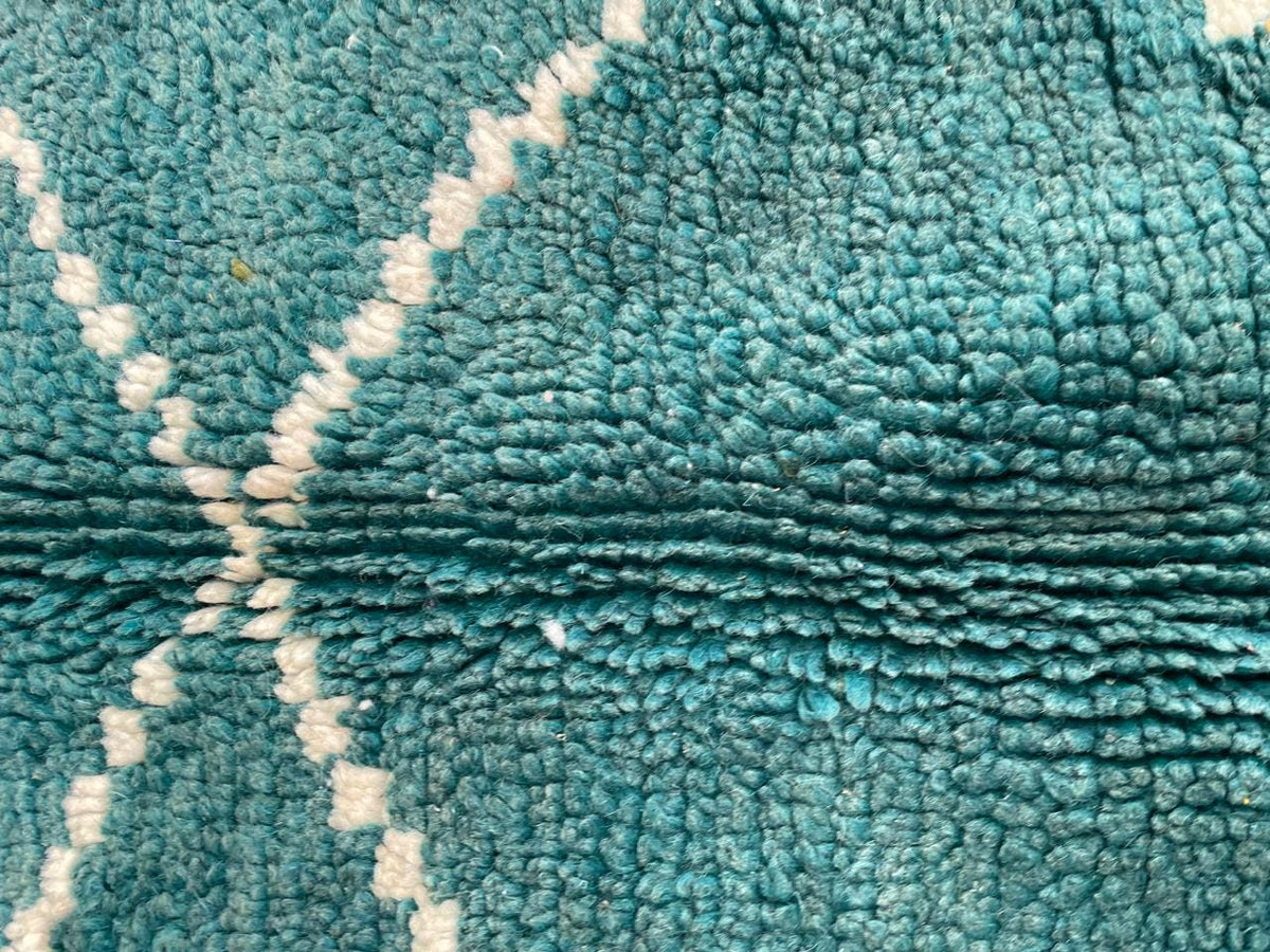 Detail close-up of a green turquoise rug featuring a textured surface and white stitching on a varied background.
