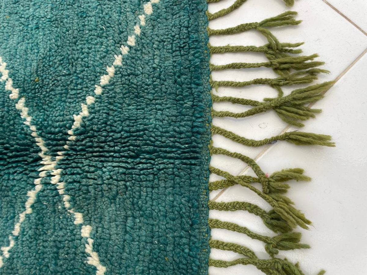 Detail close-up of a green wool rug with fringe edges and a textured surface on a white background.