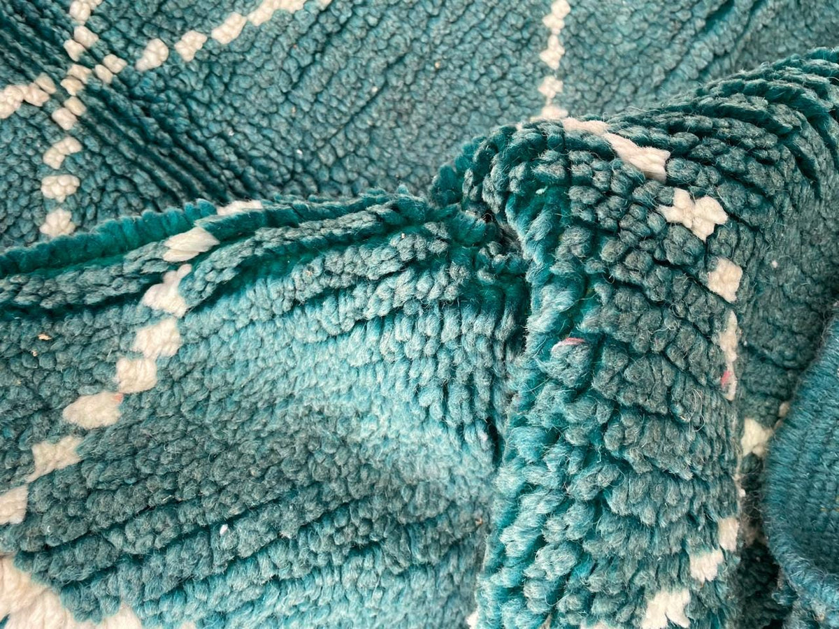 Detail close-up of a green turquoise rug displaying a diamond pattern and folded edge on a varied background.