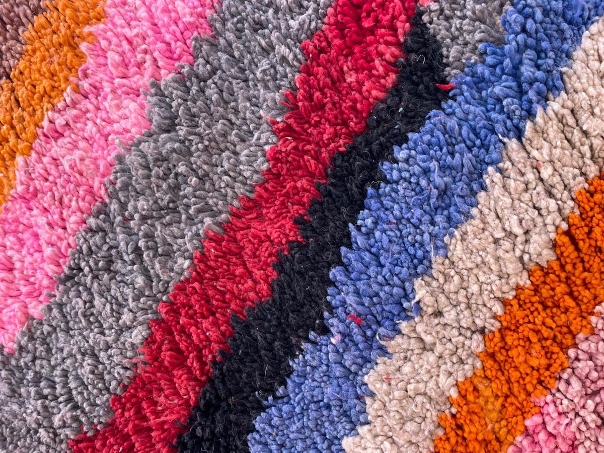 Close-up detail of the textured fibers of a colorful rug with stripes in pink, gray, red, and orange.
