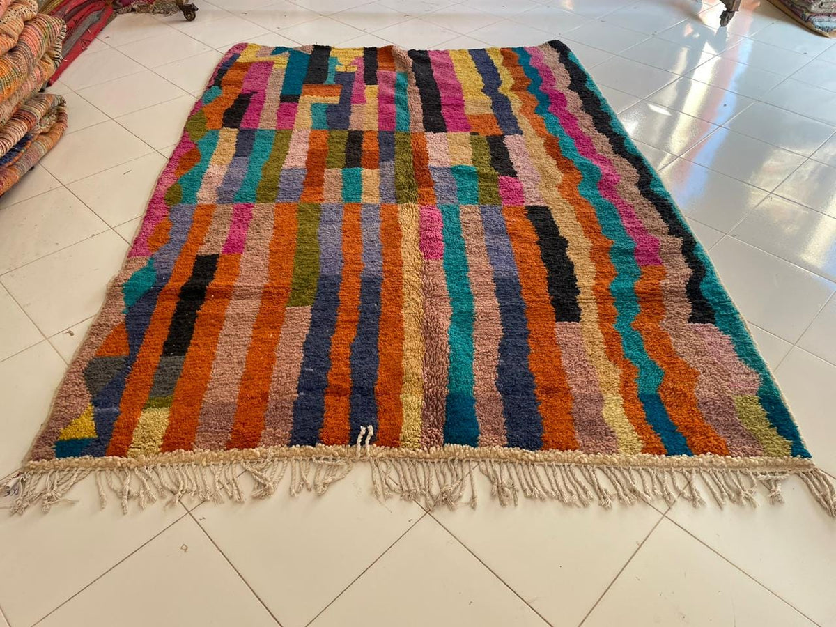 Top view of a colorful Moroccan rug featuring a striped pattern and fringed edges in pink, blue, and brown.