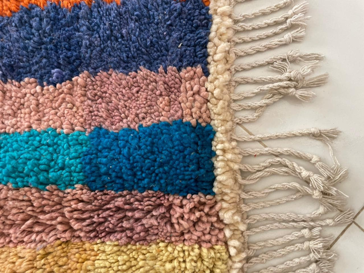 Detail shot of a colorful Moroccan rug with a striped pattern and fringe in blue, pink, orange, and yellow.