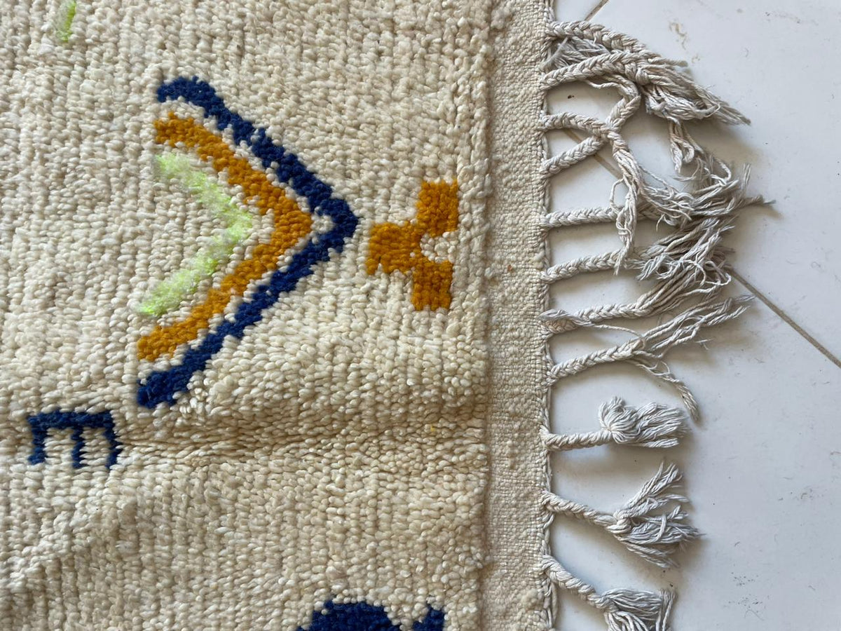 Detail shot of a handmade rug with fringe edges and geometric patterns on a white background.